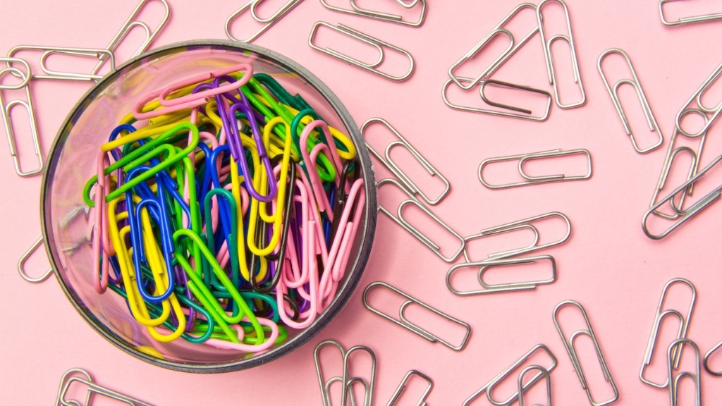 A bowl filled with colorful paper clips is surrounded by silver paper clips scattered on a pink background.