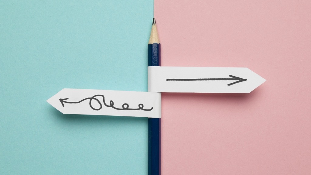 A pencil stands vertically between a blue and a pink background, with two arrow signs pointing in opposite directions—one straight and one messy and curled.