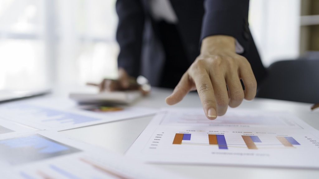 A person in a suit is pointing at a paper with bar charts on a desk covered with documents.