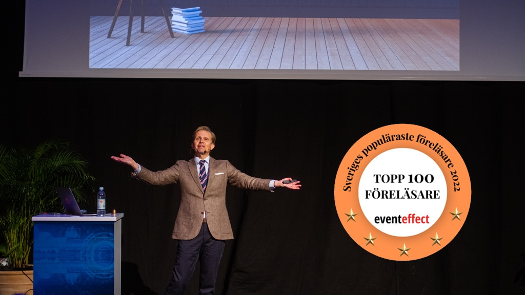 En man i brun kavaj står på ett podium och sträcker ut armarna generöst under det att han föreläser, bredvid ett emblem med texten "Topp 100 föreläsare".
