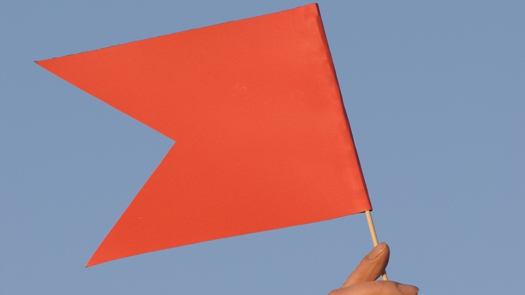 Röd pappersflagga hållen av två fingrar, mot en blå himmel.