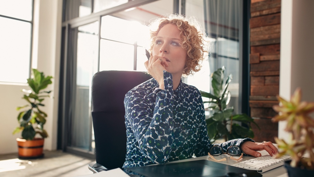 A thoughtful woman is sitting at her desk with her chin resting on her hand, looking out of the window.