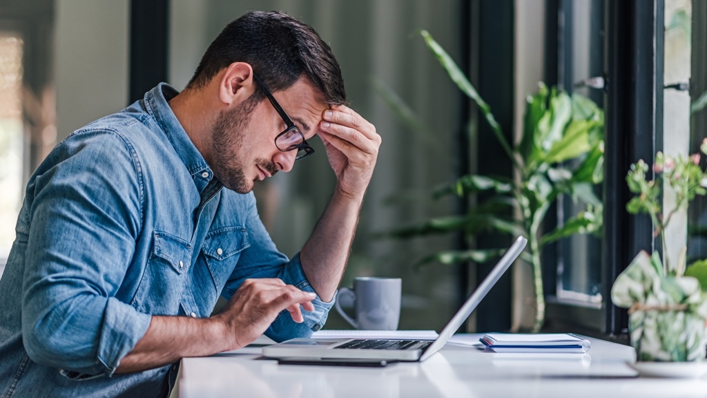 A man wearing glasses and a denim shirt sits at a desk, looking frustrated while working on his laptop, with plants and a notebook nearby by the window
