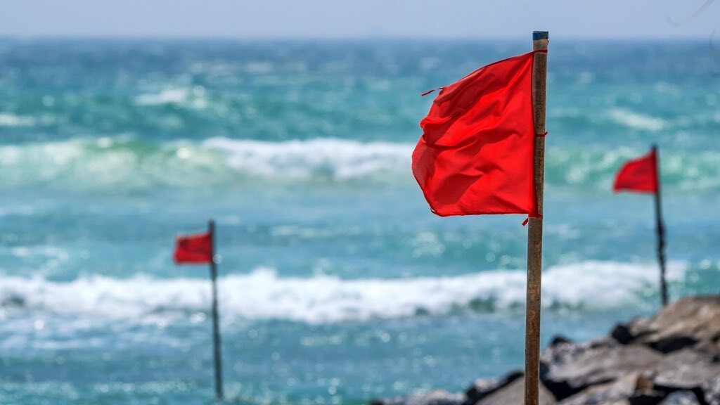 Tre röda varningsflaggor på en strand vid ett stormigt hav.