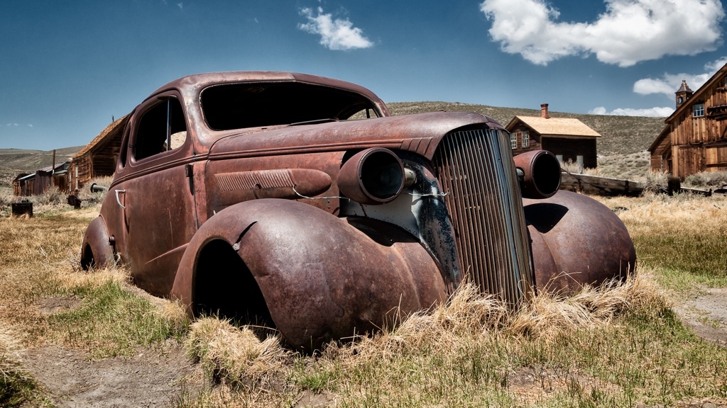 A rusty car wreck protrudes from the ground in an American ghost town on a sunny, warm day.