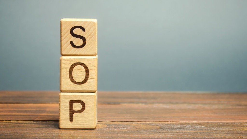 Three wooden blocks stacked vertically spell out the letters “SOP,” commonly referring to Standard Operating Procedure, on a wooden surface with a blue-gray background.