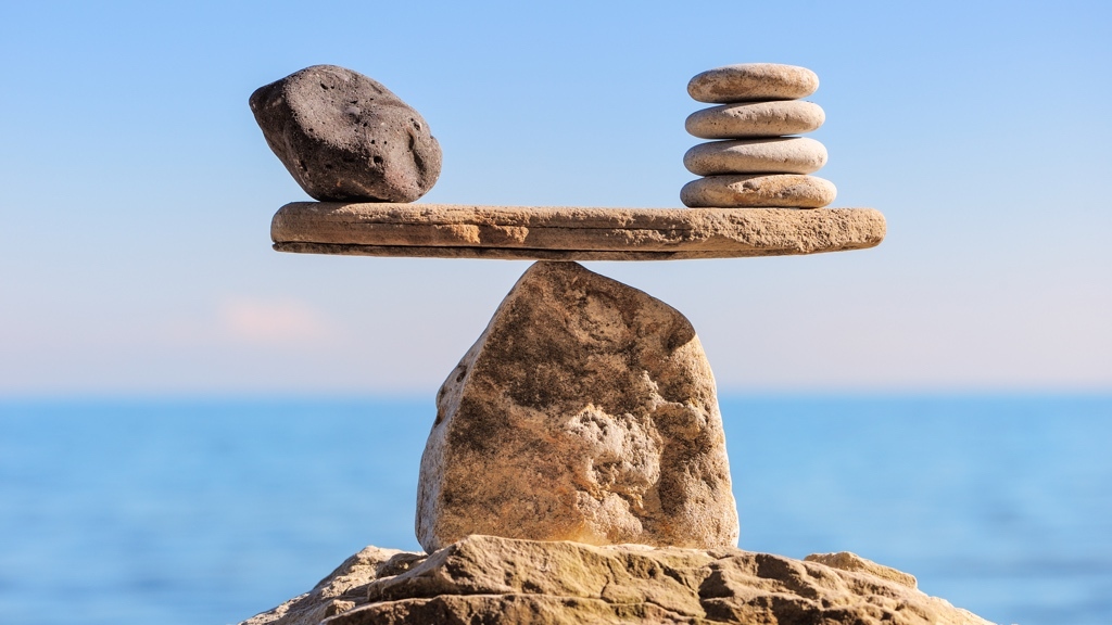 A single dark stone and a stack of five lighter stones are perfectly balanced on a plank resting atop a pointed rock, with a calm sea and clear sky in the background.