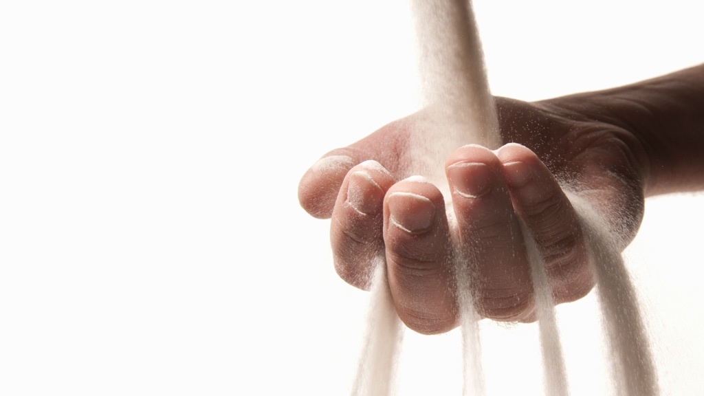 Sand rinner mellan fingrarna på en hand som sträcks in från höger i ett vitt rum.