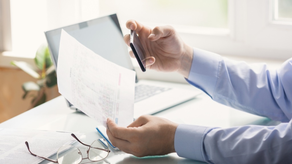A person is sitting at a desk holding a printed document in one hand and a pen in the other, with a laptop and glasses nearby.