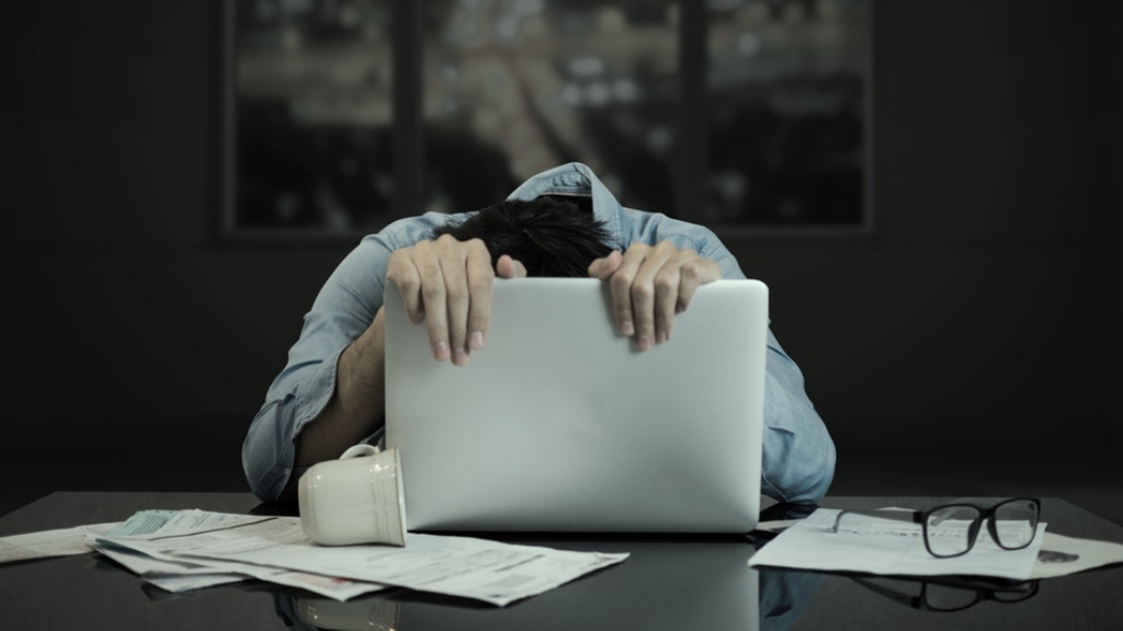 En person ligger med huvudet mot sin laptop, omgiven av papper, en mugg och glasögon, vilket antyder stress eller utmattning på grund av överarbete.