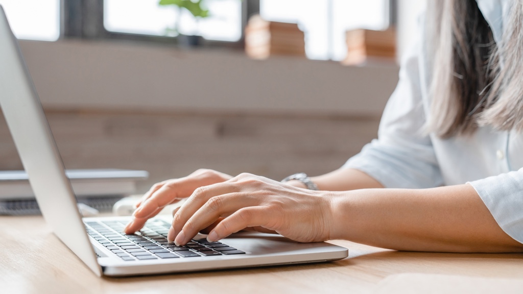 A woman's hands typing on a laptop keyboard in a light office setting. She wears a light blue blouse.