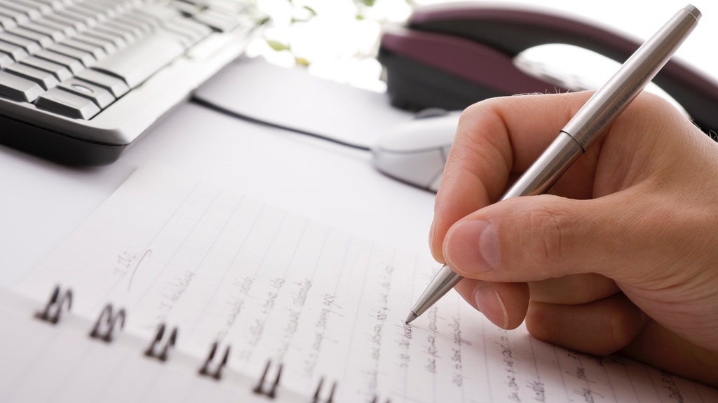 A hand is writing on a lined notepad with a pen, with a keyboard and computer mouse in the background.