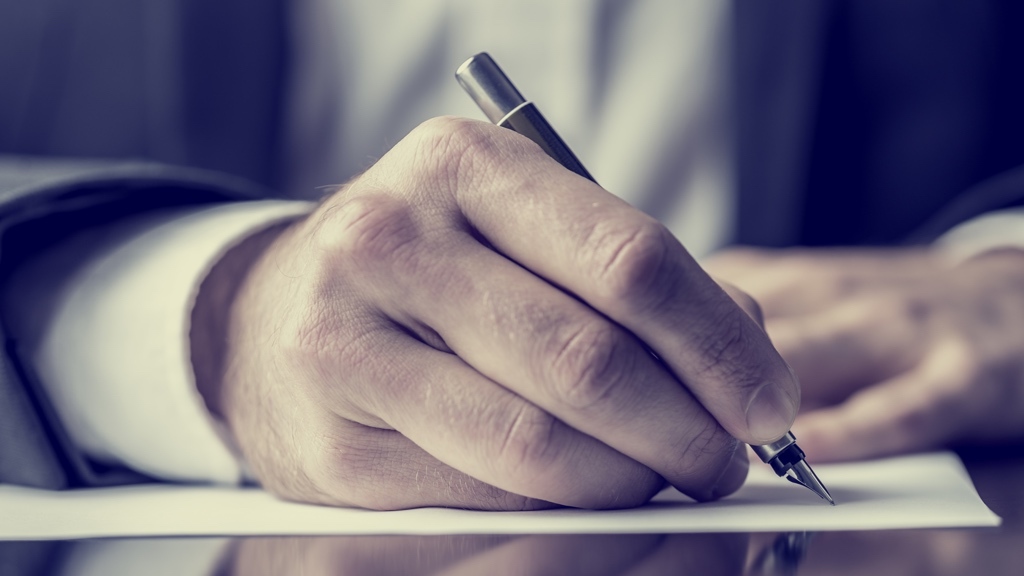 Close-up of a right hand holding a pen writing on a sheet of white paper on a desk.