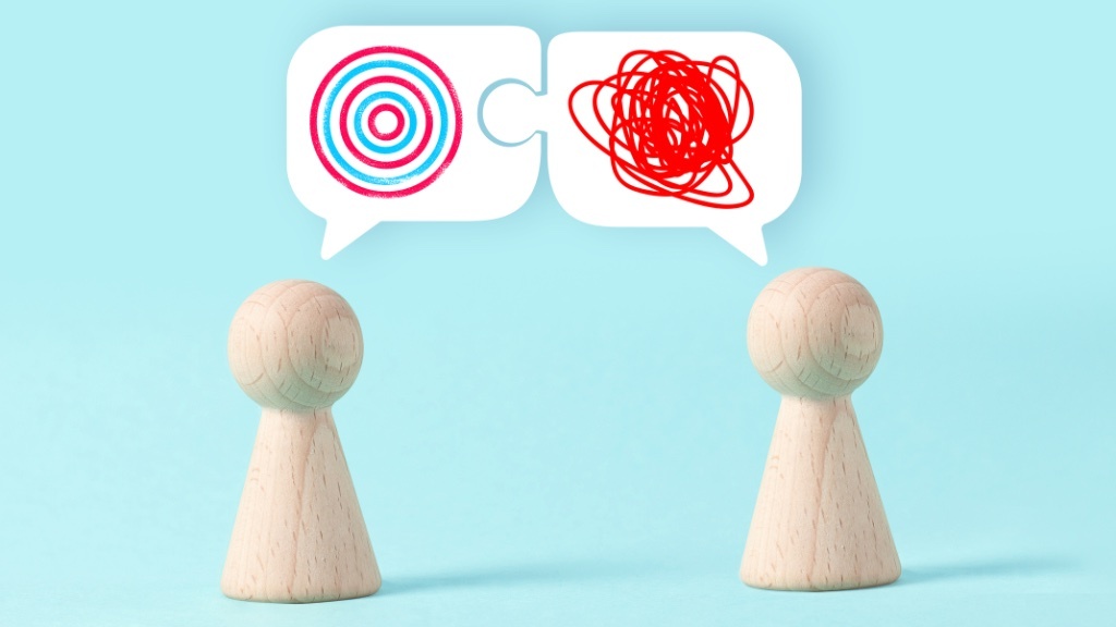 Two wooden figurines face each other with speech bubbles above them, one containing concentric circles and the other a chaotic red scribble, symbolizing a communication mismatch or misunderstanding.