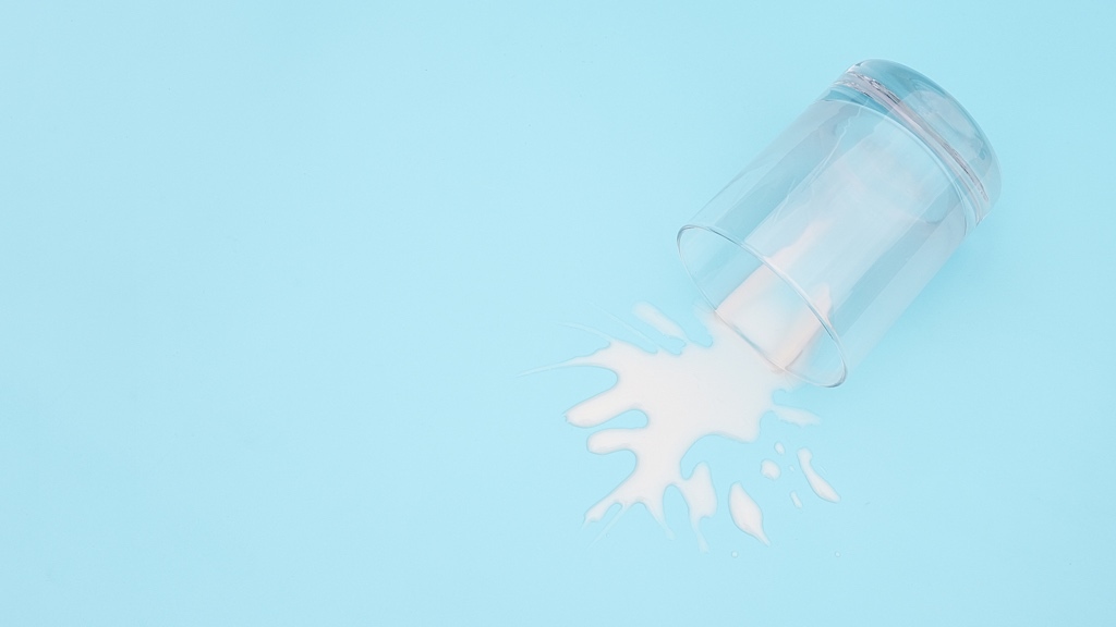 A clear glass has tipped over, spilling milk onto a light blue surface.