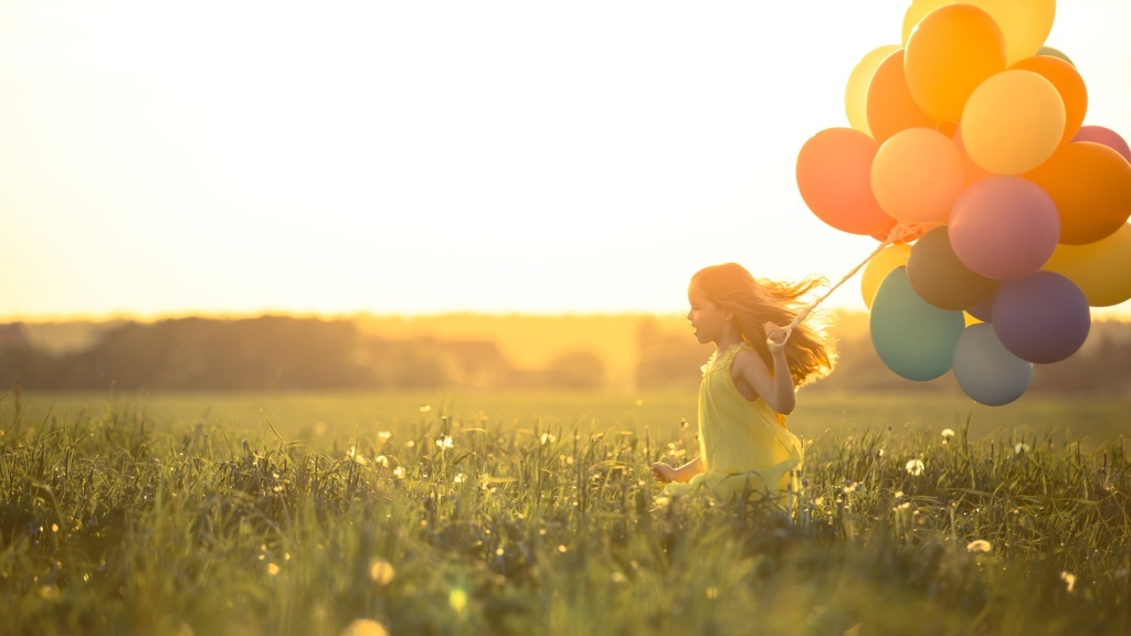 En liten flicka springer i motljus på en blomstrande sommaräng. I handen håller hon ett stort knippe färglada ballonger.