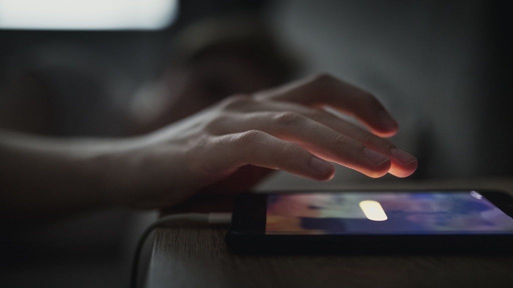 En hand sträcker sig i mörkret för att stänga av larmet på en smartphone som ligger på ett nattduksbord.