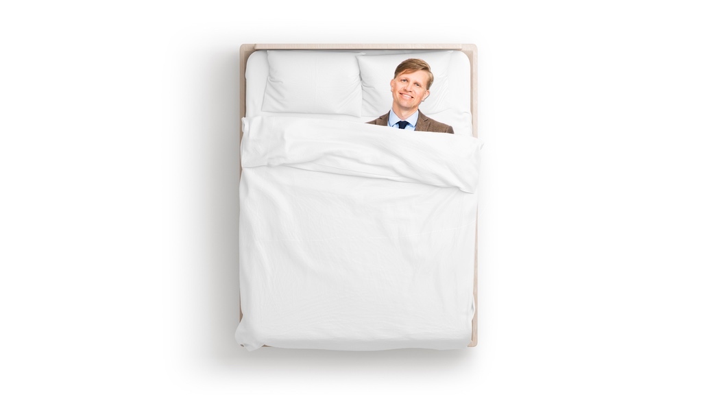 En leende man i brun kavaj och mörkblå slips ligger i en nybäddad säng.