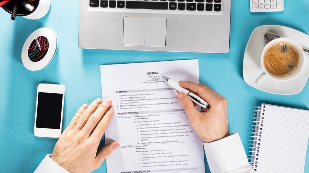 A person is reviewing a printed résumé at a neatly organized desk with a laptop, coffee, smartphone, notepad, and office supplies.
