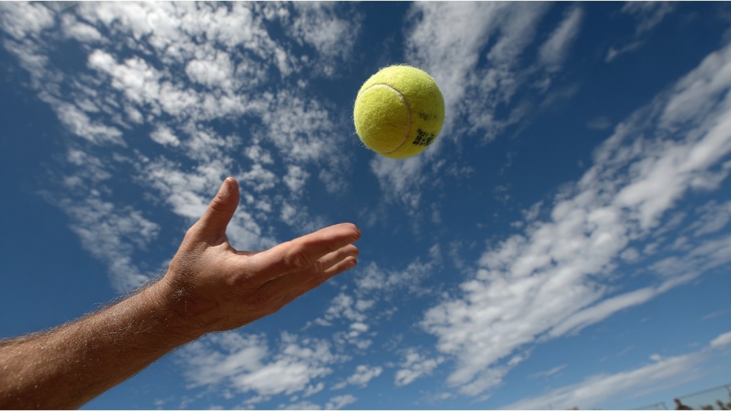 En hand kastar upp en gul tennisboll i luften mot en blå himmel med spridda moln.
