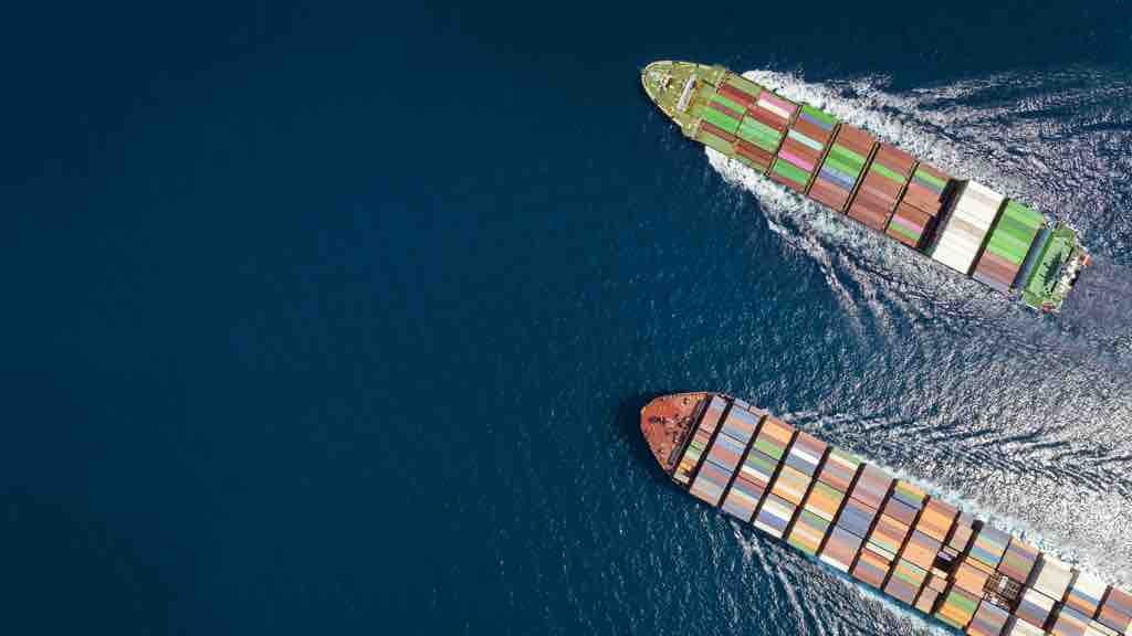 Two large container ships travel across the open sea, leaving white trails in the water as seen from above.