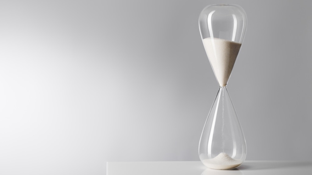 A slender hour glass entirely in glass on a white table top.