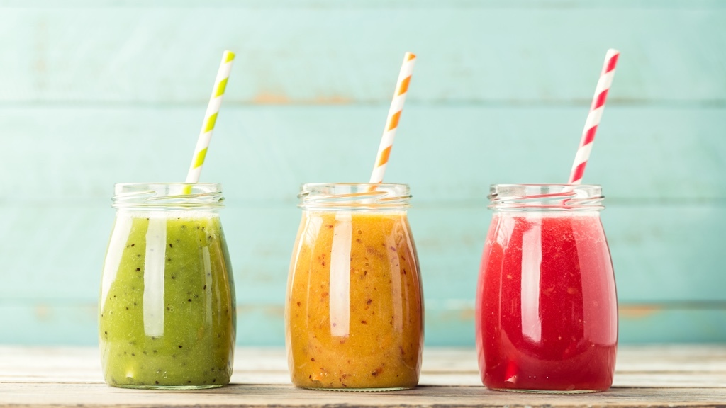 Three glass jars filled with colorful smoothies—green, orange, and red—each with a striped paper straw, are placed in a row against a light blue wooden background.