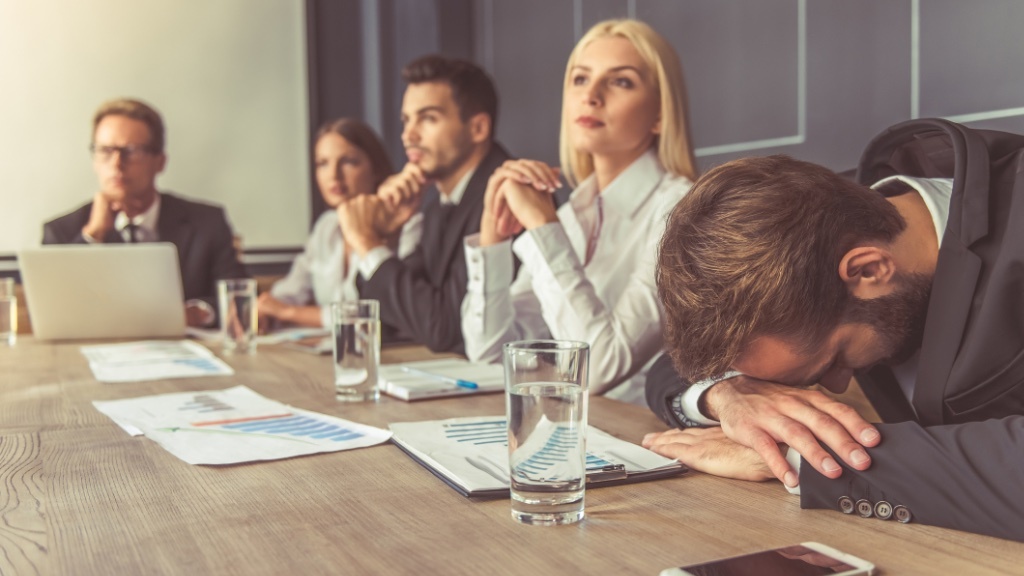 One person has fallen asleep at a long, serious business meeting while the others try to stay attentive.