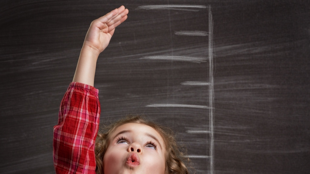 A child in a red plaid shirt is reaching up to chalk height tallying marks on a blackboard.