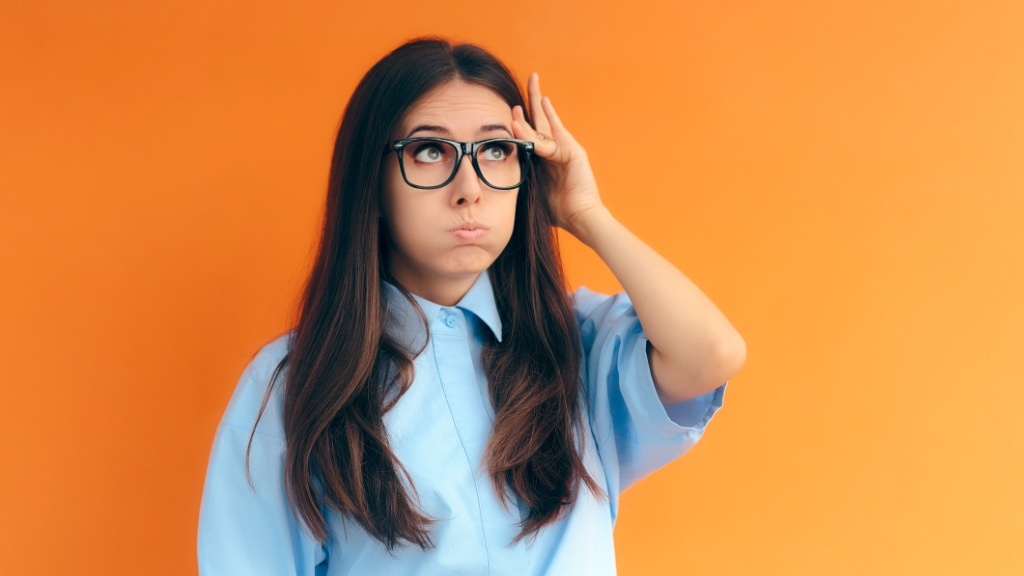 A young woman with long hair and glasses is puffing out her cheeks and looking up thoughtfully, standing against a bright orange background.