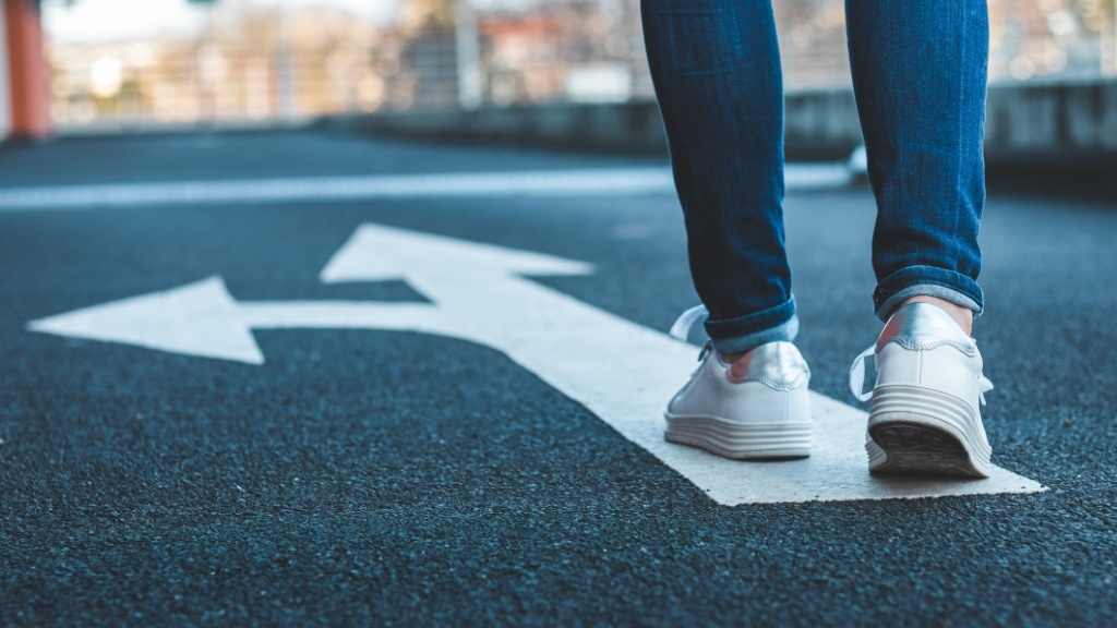 A person wearing white sneakers and jeans is walking along a road with large painted arrows pointing left and right, symbolizing a choice or decision ahead.