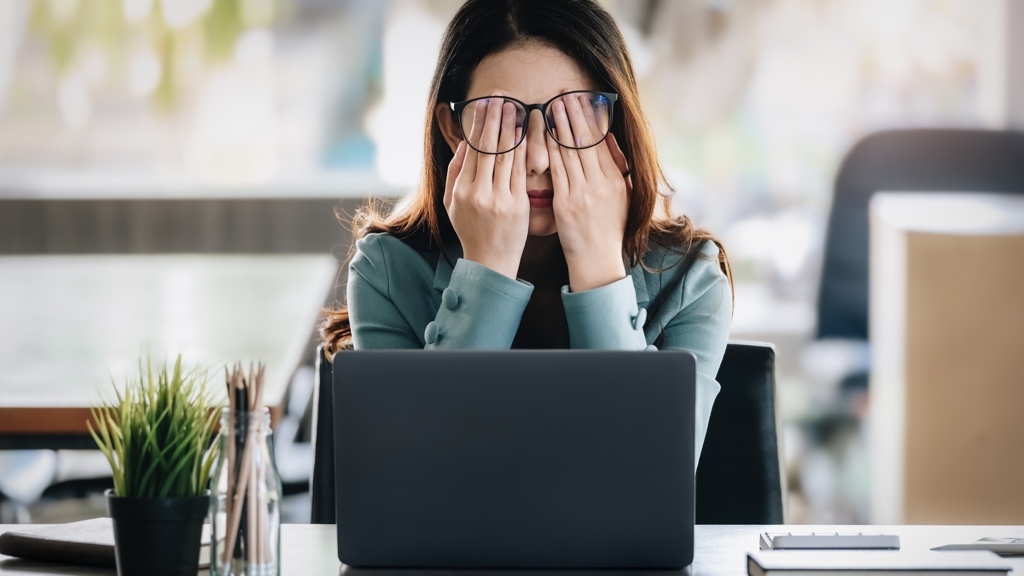 A woman sitting at her desk covers her face with her hands in frustration or exhaustion, with an open laptop in front of her in a bright office setting.