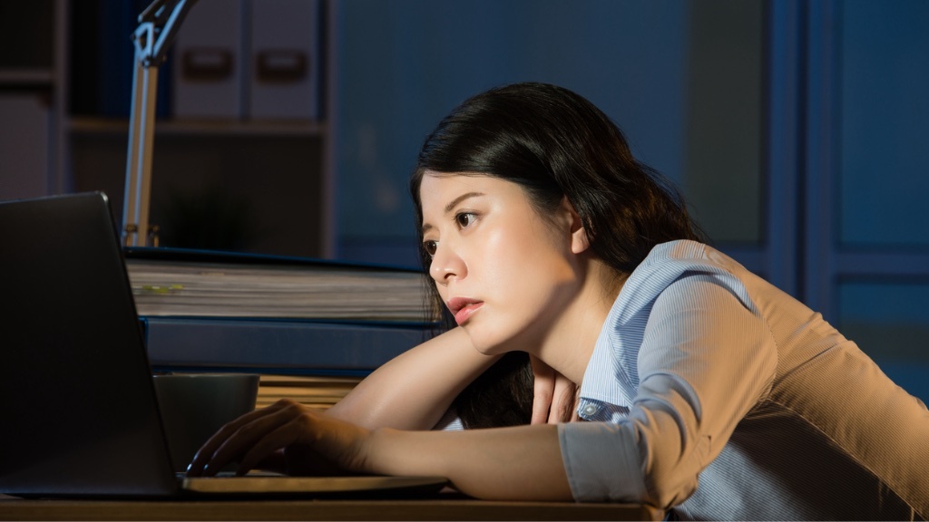 A woman appears tired and leans on her desk while staring at her laptop late at night, surrounded by files and office supplies.