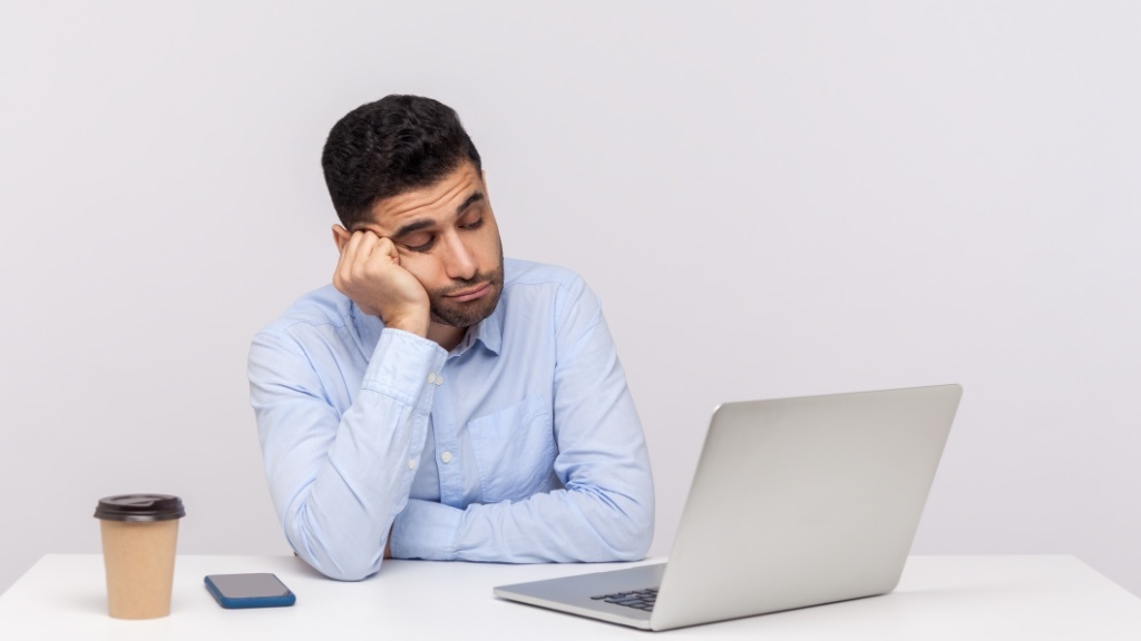 A tired-looking man in a blue shirt rests his head on his hand while staring at a laptop, with a coffee cup and smartphone beside him on the desk.