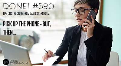 A professional woman in a black blazer is multitasking, speaking on the phone while writing notes and looking at a laptop.