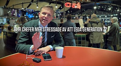 En man i tweedkavaj och randig slips sitter vid ett rött bord på ett livligt café, gestikulerar medan han talar, med en kaffekopp, smartphone och glasögon framför sig, medan en fråga om stress vid ledarskap av Generation Z visas på bilden.