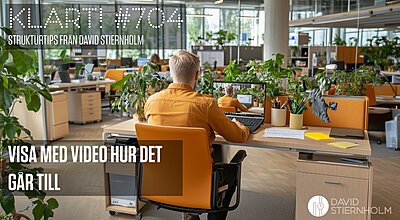 En man i en orange skjorta sitter vid ett modernt skrivbord med en orange stol, arbetar på sin laptop och är omgiven av gröna växter i en kontorsmiljö med öppet landskap.