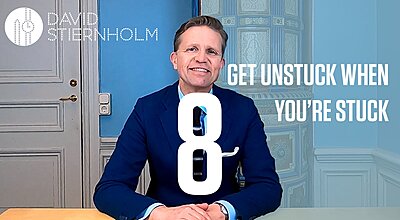 A man in a blue suit is smiling at a desk with the text “GET UNSTUCK WHEN YOU’RE STUCK” and a large number 8 in front of him.