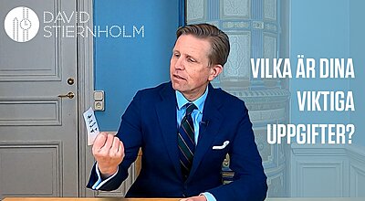En man i kostym sitter vid ett bord och tittar fundersamt på en liten lapp han håller i, med texten “VILKA ÄR DINA VIKTIGA UPPGIFTER?” och “DAVID STIERNHOLM” visad på väggen bakom honom.