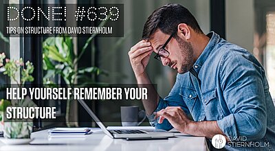 A man in a denim shirt sits at a desk, looking stressed as he focuses on his laptop with one hand on his forehead.
