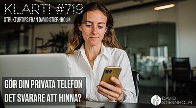 En kvinna i 30-årsåldern med långt brunt hår och vit skjorta sitter vid ett skrivbord i ett modernt kontor, tittar på sin smartphone samtidigt som hon använder sin laptop.
