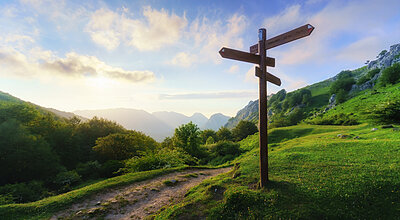 En träskylt står vid ett vägskäl i ett frodigt grönt bergslandskap under en färgstark morgonhimmel.
