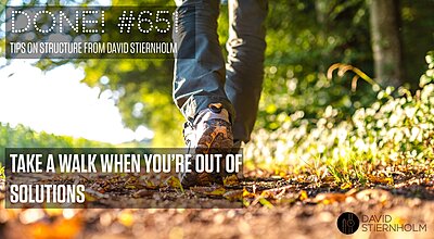A person is walking along a sunlit forest path, with the focus on their shoes and the ground covered in fallen leaves.