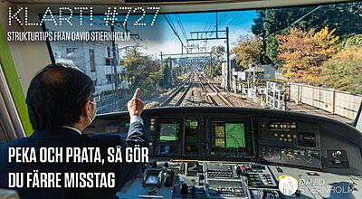 En lokförare i uniform pekar framåt medan han kör ett modernt tåg, med flera digitala kontrollpaneler framför sig och järnvägsspår som sträcker sig ut genom ett stadslandskap.