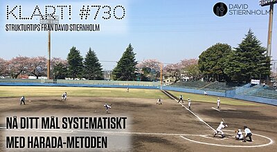 En ungdomsbasebollmatch pågår på en grusplan, med spelare i vita dräkter och blommande körsbärsträd i bakgrunden.