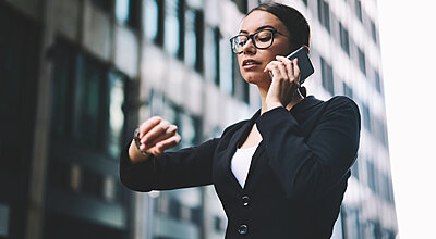 A woman in a blazer talks on her mobile phone while checking her watch outside a modern office building.