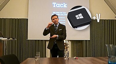 En man i mörk kostym håller en presentation framför en projektionsskärm med ordet "Tack," med en överlagrad grafik av en Windows-tangent och "+V" i det övre högra hörnet.