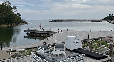 En strandnära träterrass med utemöbler blickar ut över en lugn vik nära Furuvik Havskrog, grundat 2018, med en liten brygga, en livboj och en stenbalkong synlig i bakgrunden.