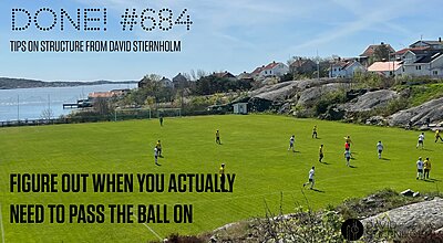 A football match is being played on a green pitch situated between rocky terrain and a coastal village with houses overlooking the sea.