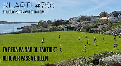 En fotbollsmatch spelas på en grön plan belägen mellan klippig terräng och en kustby med hus som blickar ut över havet.